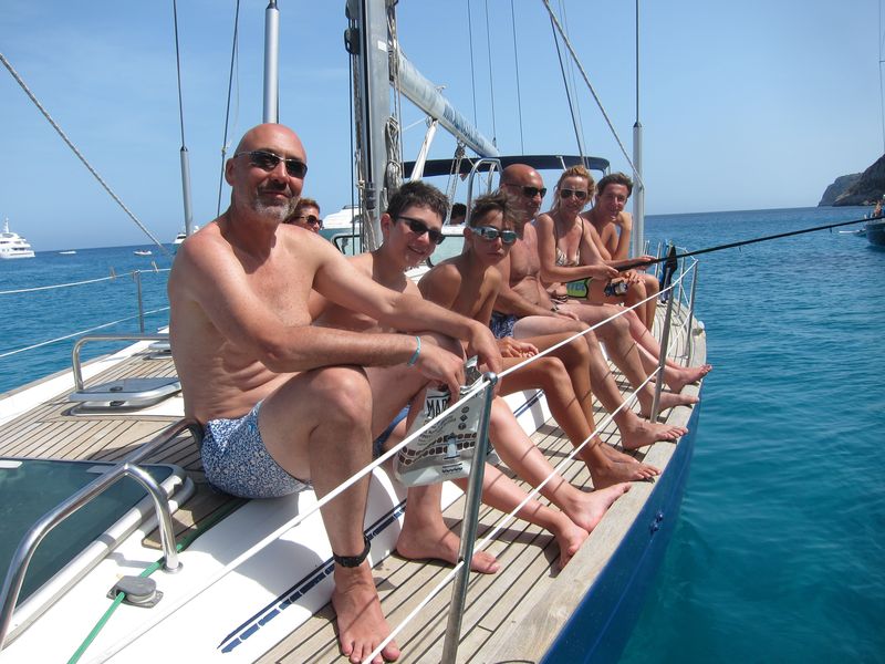 Una familia con niños disfruta de sus vacaciones de alquiler veleros Ibiza. Aparecen sentados en la banda de babor de un velero. Una mujer está pescando con una caña de pescar.